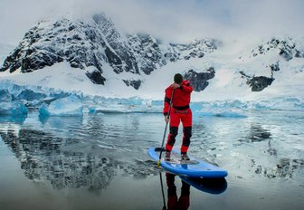 A man in a drysuit on a paddleboard in the icy waters of Antarctica with snowy peaks in the background