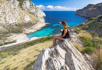 girl sits atop a rock looking out over a white sand, idyllic beach in Mallorca
