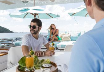 male guest is served a cocktail at the bar while female guest sits in lounging area on sundeck aboard luxury yacht TOUCH
