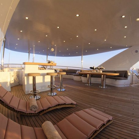 Overview of the sheltered section of the sun deck onboard charter yacht GRANDE AMORE, sun loungers in the foreground with a bar and stools in the background. 