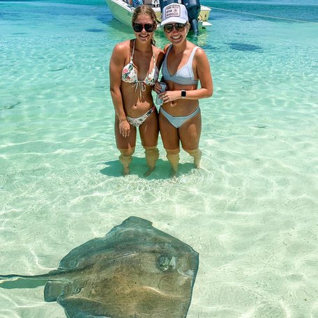 Girls in bikinis standing with a stingray in the water while a sailing boat is anchored behind them 