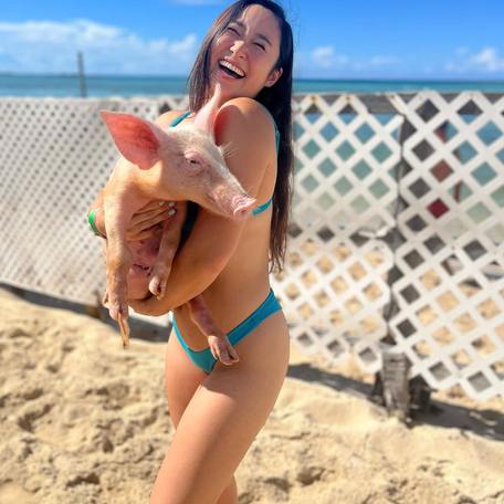 Girl in bikini standing on the sand holding a pig 