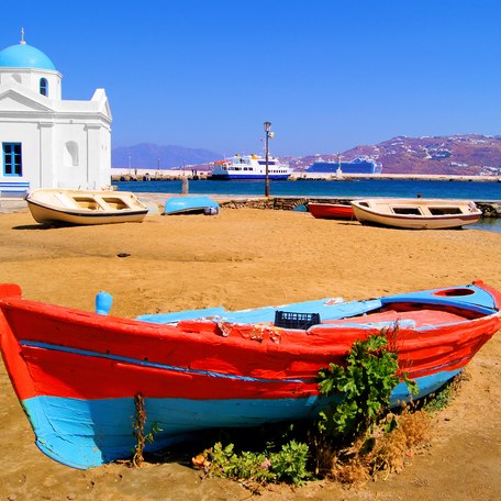 A small red and blue boat sits on a sandy beach during a Mykonos yacht charter with a small church in the background