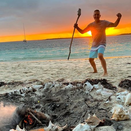 Man standing holding wooden staff by a dying beach bonfire at sunset 