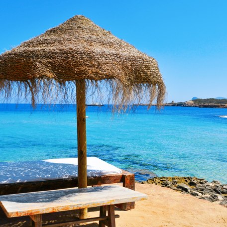 Detail of a relaxing area in a Cala Conta beach in Ibiza Island, Spain, with a comfortable sunlounger and a rustic umbrella made of natural fibers