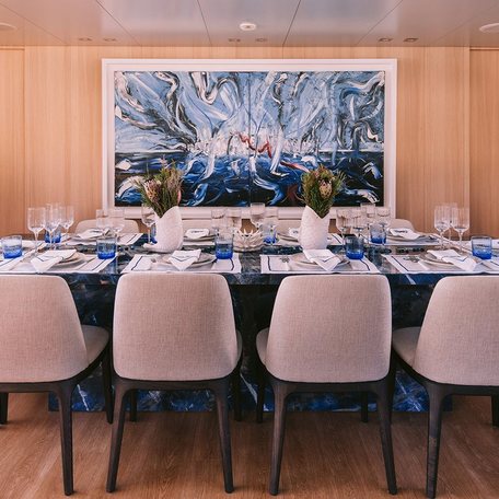 Overview of an interior dining table onboard charter yacht ISLANDER II, ten seats surround a rectangular table with a painting on the wall behind. 