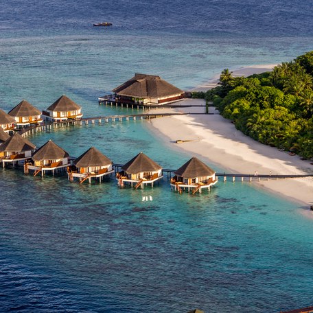 Water Huts and Beach Villas. Crystal blue sea and white sandy beach with green flora. Reefs also visible in the lagoon.