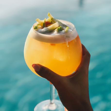 Close-up of hand holding orange cocktail in front of turquoise blue water 