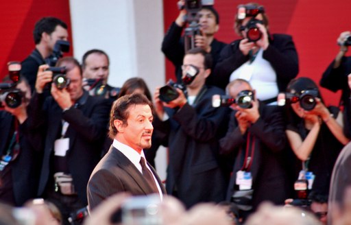 A leading man faces cameras on the red carpet wearing a black suite and red tie at the Venice Film Festival