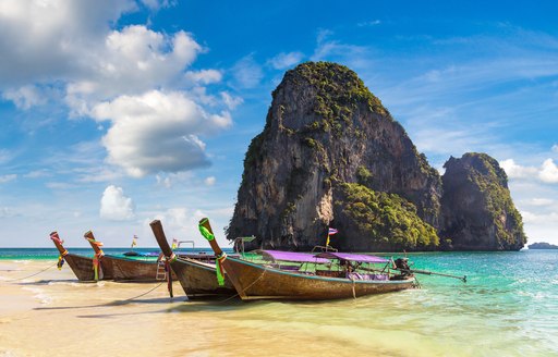 Long tailed boats are pulled up onto a golden beach lapped by green waters with a limestone rocky outcrop in the distance