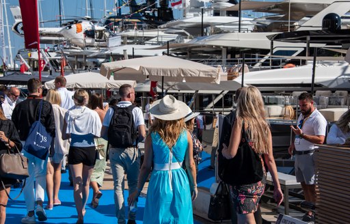 Visitors to the Monaco Yacht Show walking along a blue carpet, with superyachts berthed to the right.