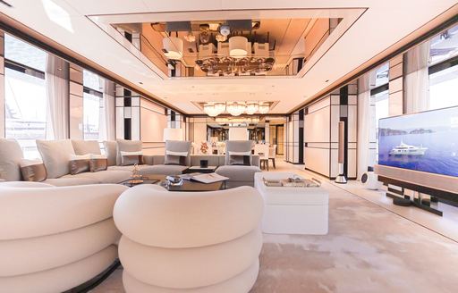 Overview of main salon onboard charter yacht RELIANCE, with large windows, plush cream seating and a large TV to starboard