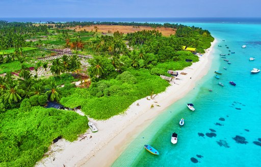 Coastline of a Maldivian island. Lush green foliage bordered by white sands overlooks anchored tenders and clear waters.