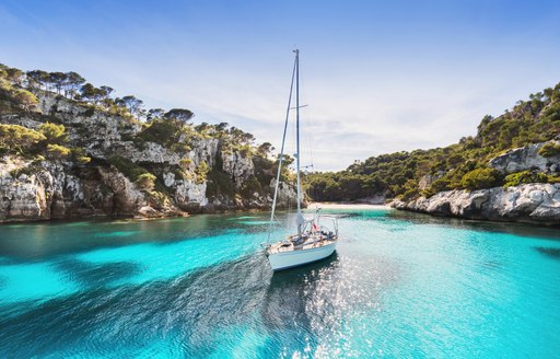 little sailing boat sits in small cove in the balearic islands, surrounded by rocks and forest