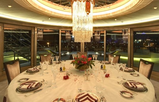 Formal dining arrangement onboard yacht charter AMARYLLIS. Round table ornately displayed with hanging chandelier directly above, surrounded by extensive windows.