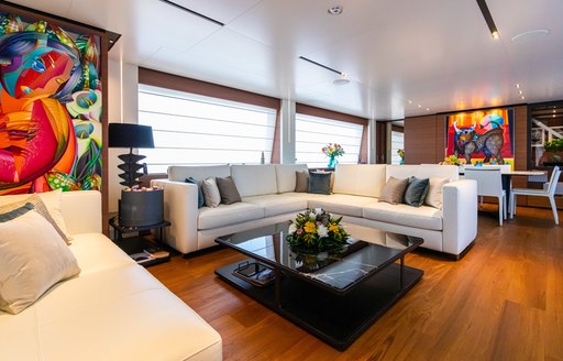 Main salon onboard Ferretti yacht charter 30m PENELOPE, white corner sofa around a black coffee table, coloured paintings on surrounding walls