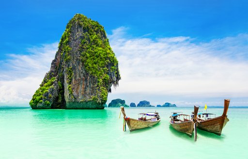 long tailed boats are pulled up onto a golden beach lapped by green waters with a limestone rocky outcrop in the distance