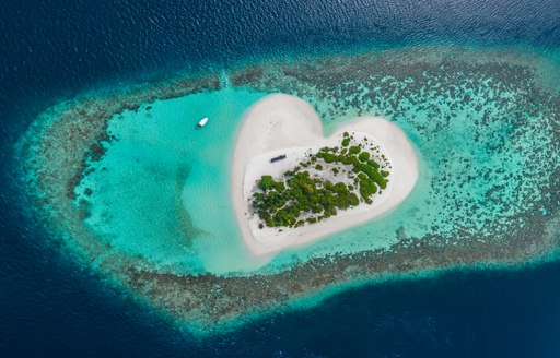 Aerial view of heart shaped island in the Maldives, surrounded by a coral reef separating azure and sapphire waters.