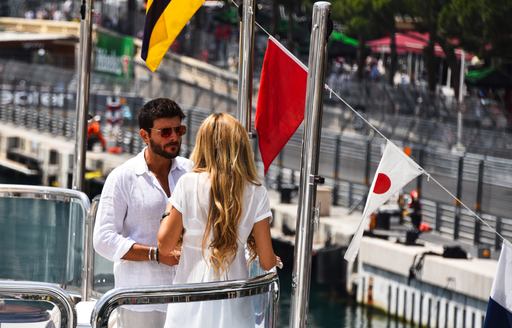 Man and woman stand on bow of a luxury yacht having a chat, with race circuit visible in background