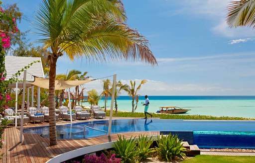 man walks next to infinity pool on thanda island. Sun loungers are lined up, along with palm trees and bright blue ocean in background