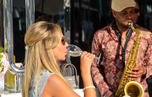 Woman enjoying champane on board superyacht with saxophonist in background at Monaco Yacht Show 2018