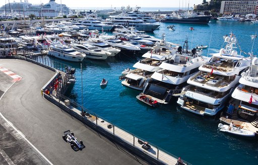 Row of superyachts moored at Monaco Grand Prix, F1 car speeding past on track adjacent to marina.