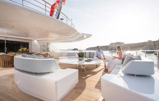 Couple sitting on aft deck of superyacht O'PARI on light colored furnishing in sun