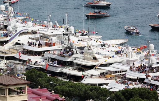 Row of superyachts moored at Monaco Grand Prix, flocks of guests on each yacht enjoying party atmosphere.