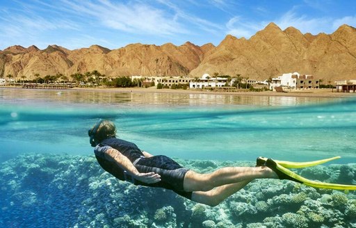 Charter guest snorkelling in the Red Sea, glimpse of corals under water and old settlement against mountainous terrain in background