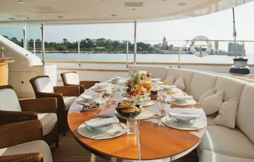 Dining set up onboard Parsifal III, external rounded sofa and large dining table accompanied by four armchairs, overlooking sea and distant palms