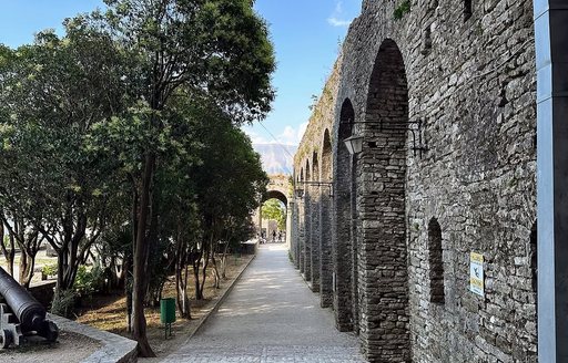 Stone pathway lined with trees alongside the fortified walls of Gjirokastra Castle in Albania, featuring historic arches and a mounted cannon under a clear blue sky.