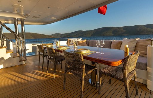 Exterior dining area on the aft main deck of M/Y IMMERSIVE, overlooking the sea and elevated terrain in the distance.