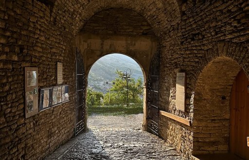 View from inside the stone archway of Gjirokastra Castle, Albania, showing cobblestone floors, historic walls, and a sunlit valley visible through the open gate.