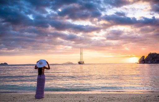 Charter guest stands on beach looking at LAMIMA yacht