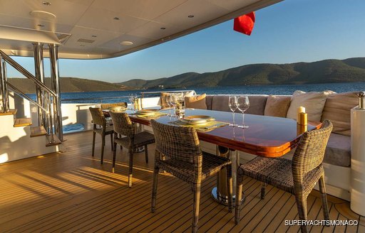 Exterior dining area on the aft main deck of M/Y IMMERSIVE, overlooking the sea and elevated terrain in the distance.