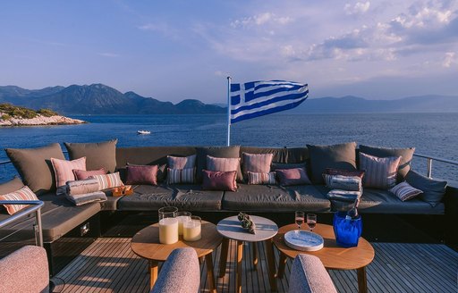 Overview of the alfresco lounge on the upper deck onboard charter yacht ISLANDER II, L shaped seating with twin coffee tables and a Greek flag in the background