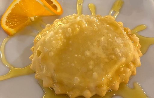 A closeup of a round Sardinian seadas pastry drizzled with honey and an orange slice on the side