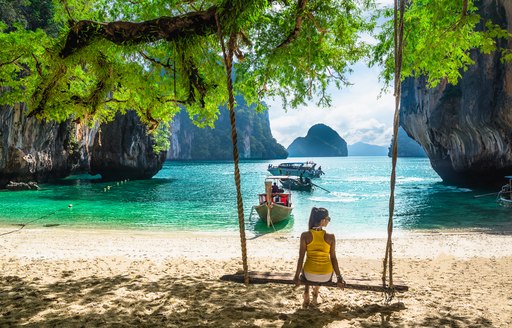 girl sitting on swing below tree on sandy beach in thailand looking out over the sea with rocks in it