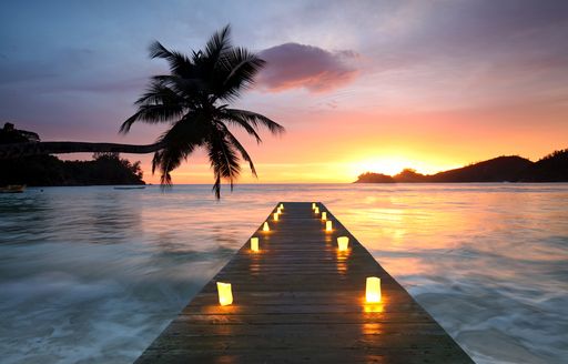 Seychelles beach overhung by a palm tree, with a wooden jetty lit up by candles at dusk