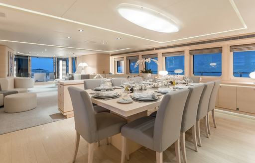 Interior dining setup onboard luxury yacht charter G3, long table surrounded by gray upholstered chairs and view of the lounge area