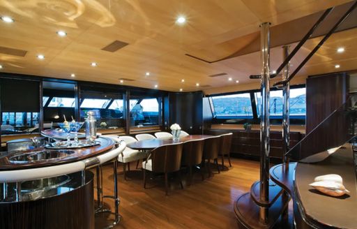 Interior dining layout onboard Parsifal III, long table adjoined by staircase and wet bar, facing wide reaching windows 