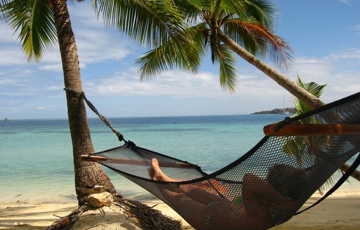 charter guests relaxes in a hammock on a sandy Fijian beach lined with palm trees