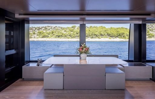 Interior dining area onboard charter yacht JICJ, central table with a bench along the back and pull out stools, with a large window in the background