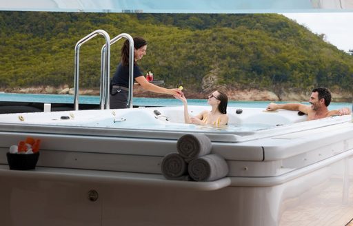 Two charter guests enjoy in a deck Jacuzzi with a crew member passing drinks onboard charter yacht ARBEMA