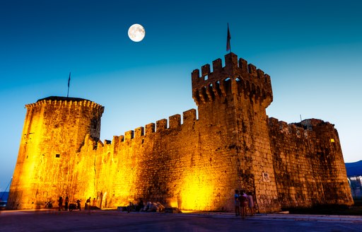 A lit up castle in Croatia at night with the moon above