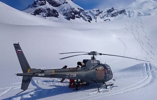A helicopter sat at the bottom of snow-capped mountains with ski tracks leading down the slope