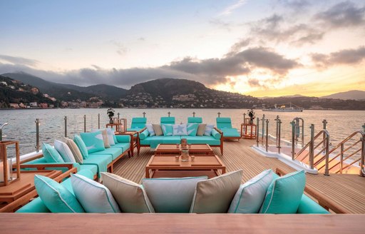 Exterior on-deck lounge area onboard charter yacht AIFER, many teal padded seats surround low coffee tables, surrounded by sea and twilight.