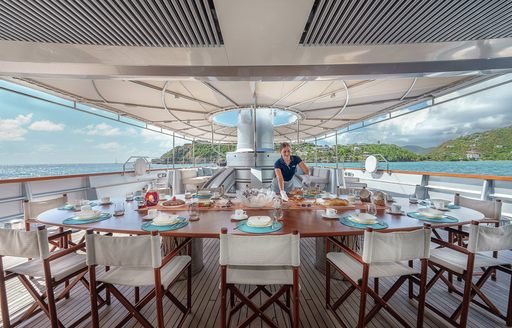 Alfresco dining set up onboard sailing yacht charter MALTESE FALCON, long table with white upholstered chairs and a crew member setting the table