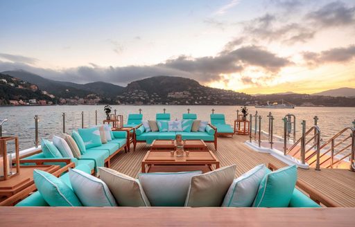 Exterior on-deck lounge area onboard charter yacht AIFER, many teal padded seats surround low coffee tables, surrounded by sea and twilight.