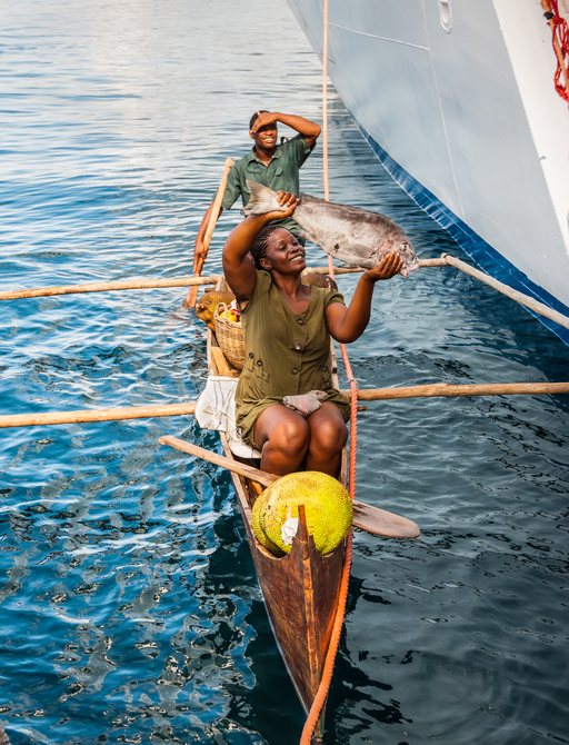 A black woman holds a fish aloft inside a small boat